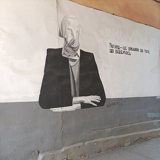 A wall mural with a person seated, dressed in black, their face is covered by a white mask. On the left is writing.